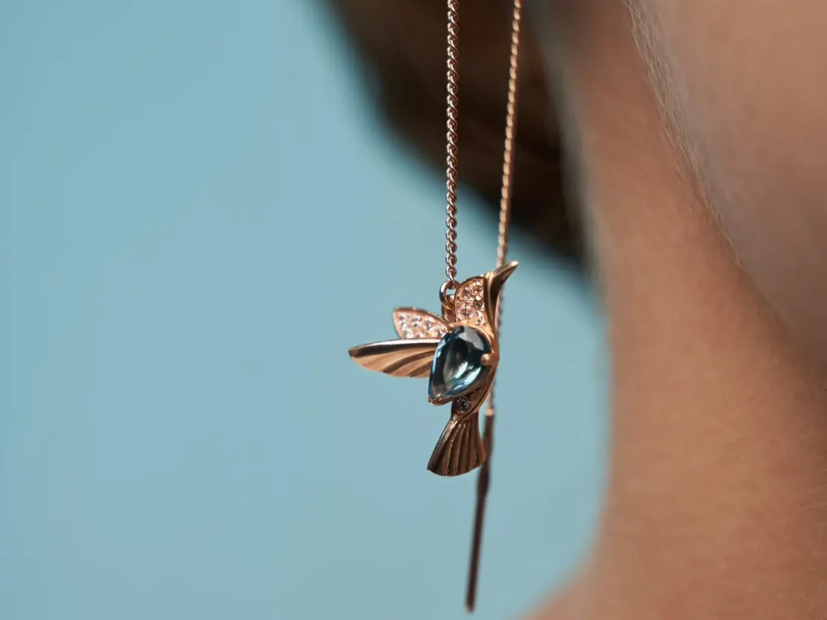 Crop of young female model presenting golden earrings in shape of bird with blue stone. Front view of incognito woman posing, isolated on blue studio background. Concept of jewelry.